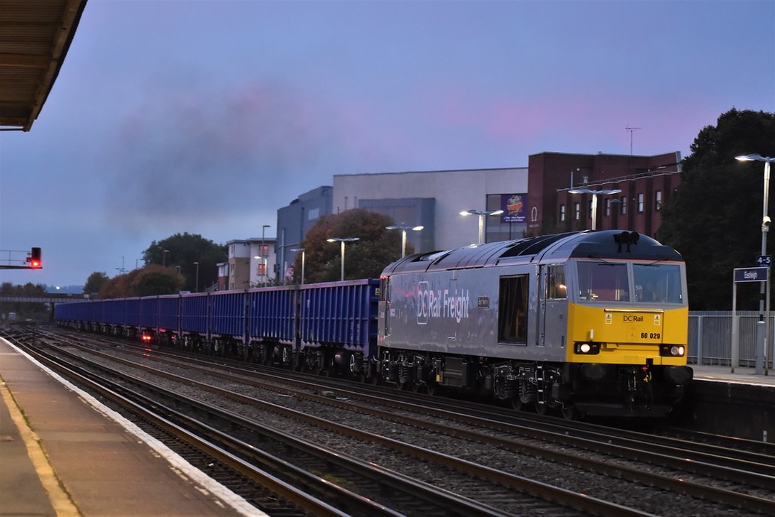 Ett modernt godståg med ett grått lok från DC Rail Freight drar en lång rad med blå containrar genom en station i skymningen.